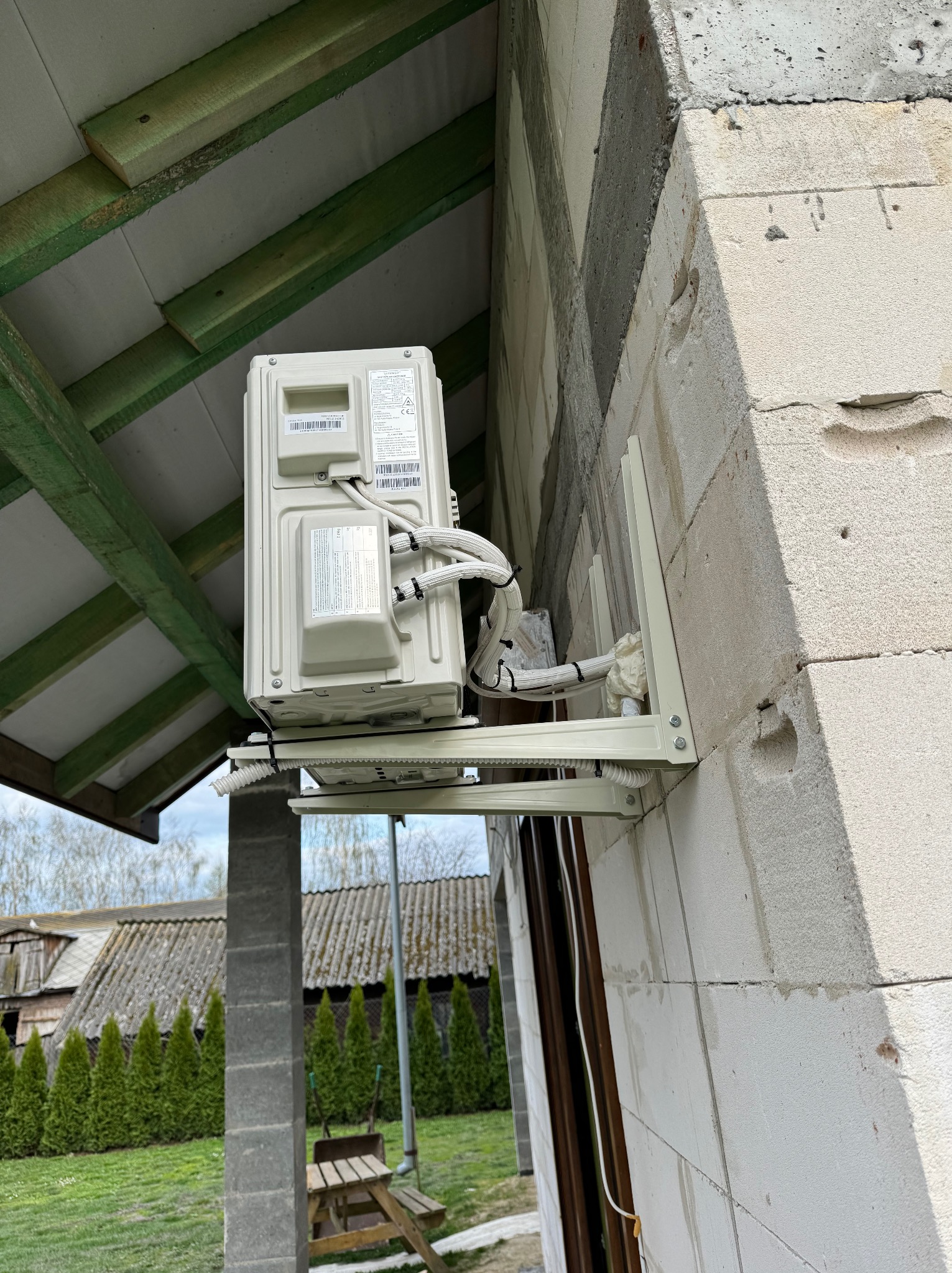 Zewnętrzna jednostka klimatyzacji zamontowana na ścianie budynku z bloczków betonowych, widoczne zabezpieczone kable i elementy mocujące.