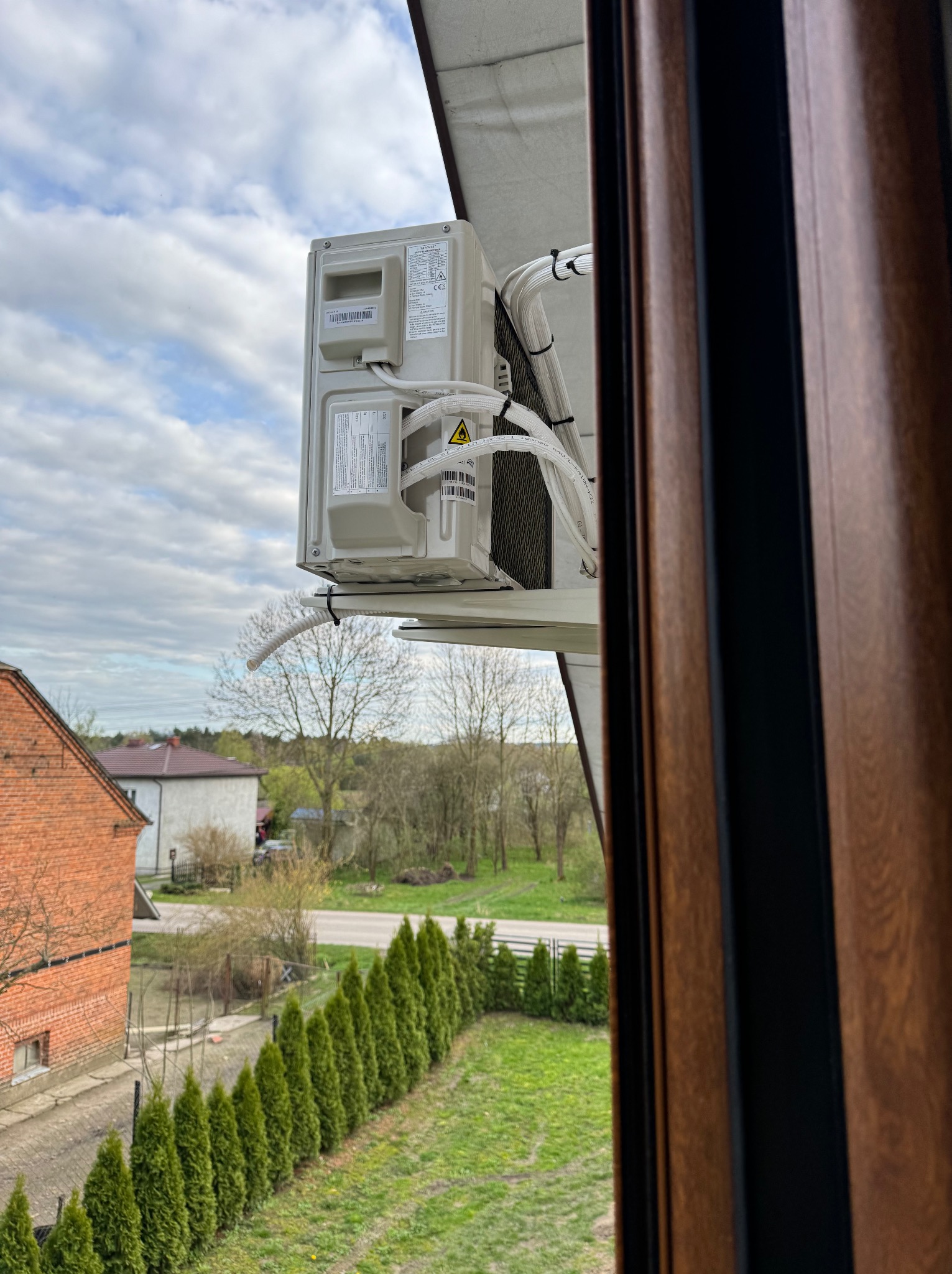 Zewnętrzna jednostka klimatyzacji zamontowana na balkonie, widoczna zza drewnianej ramy okiennej, z widokiem na ceglany budynek i rząd tuj na tle pochmurnego nieba.