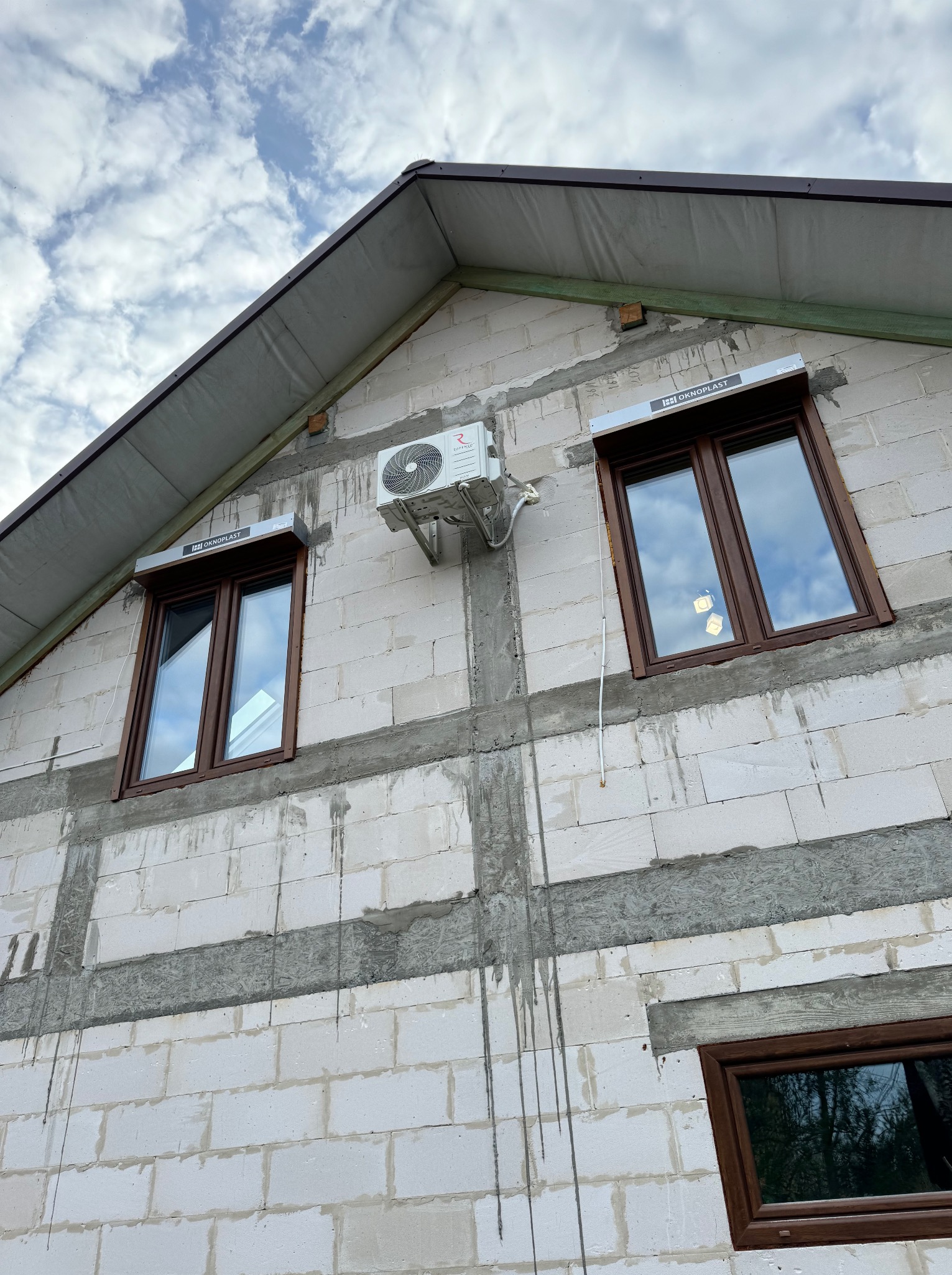 Biały budynek w trakcie budowy z zamontowaną jednostką zewnętrzną klimatyzacji obok okna z brązową ramą i roletą zewnętrzną.