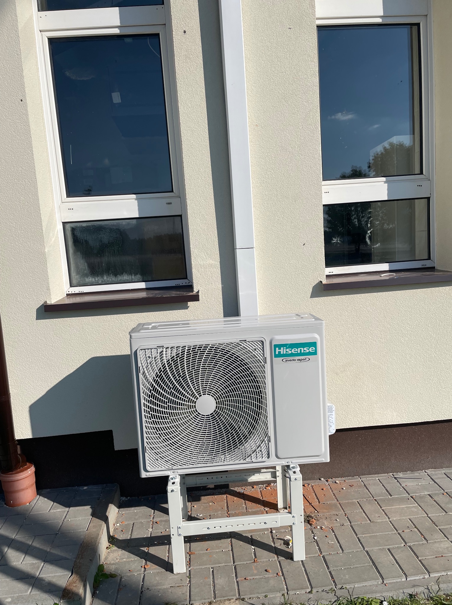 Zewnętrzna jednostka klimatyzacji Hisense zamontowana na białym stelażu przy ścianie budynku z dwoma oknami, widoczne okruchy cegieł na chodniku.