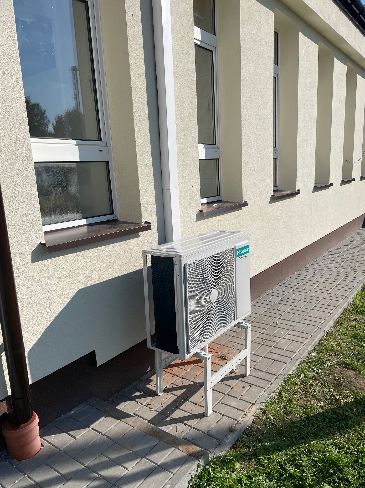 Zewnętrzna jednostka klimatyzacji Hisense zamontowana na metalowym stelażu przy ścianie budynku z rzędem okien w Lublinie, widok z poziomu chodnika z kostki brukowej.