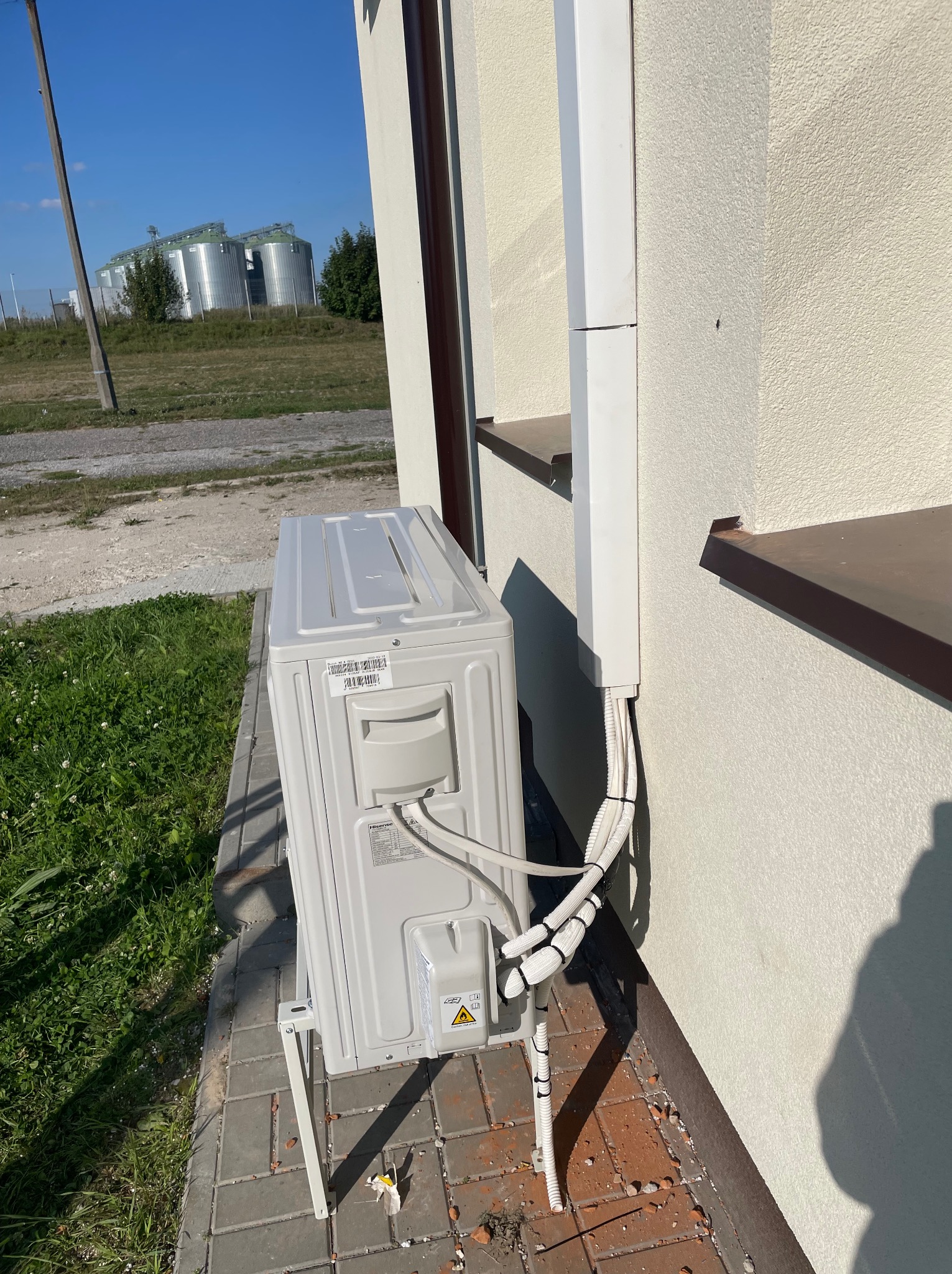 Zewnętrzna jednostka klimatyzacji typu split zamontowana na wspornikach przy ścianie budynku, z widocznymi przewodami w białej listwie i silosami zbożowymi w tle.