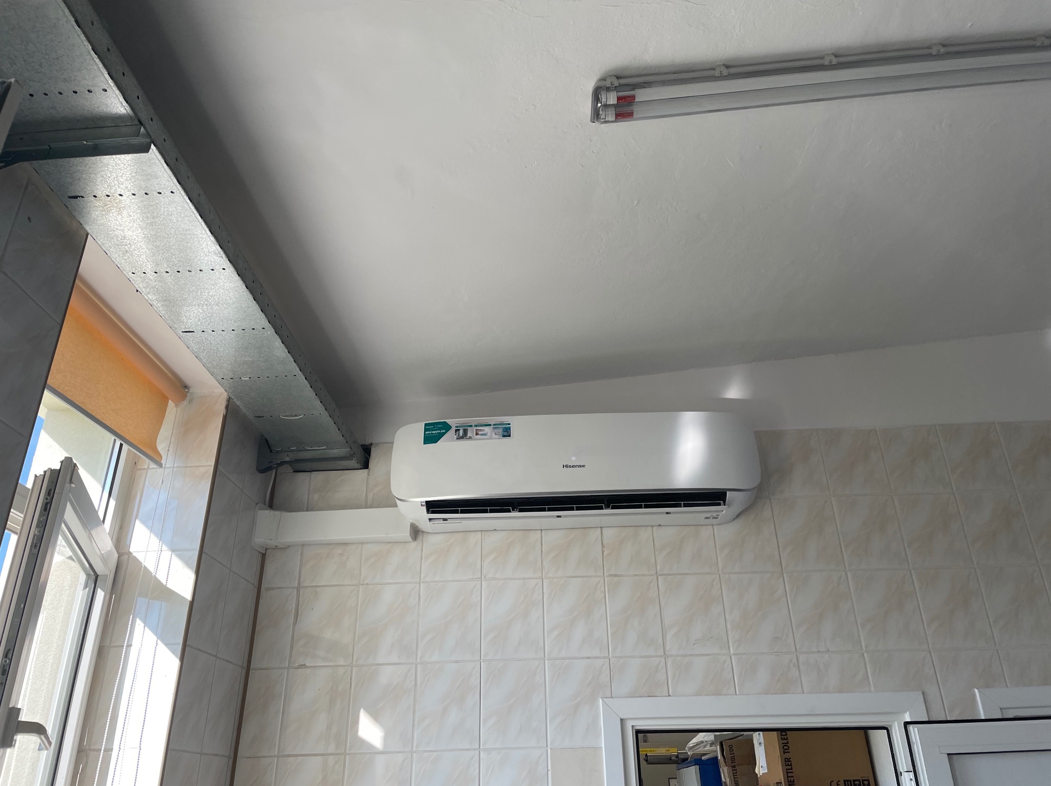 Biały klimatyzator Hisense zamontowany na kafelkowanej ścianie, widoczny kanał wentylacyjny i okno z roletą.