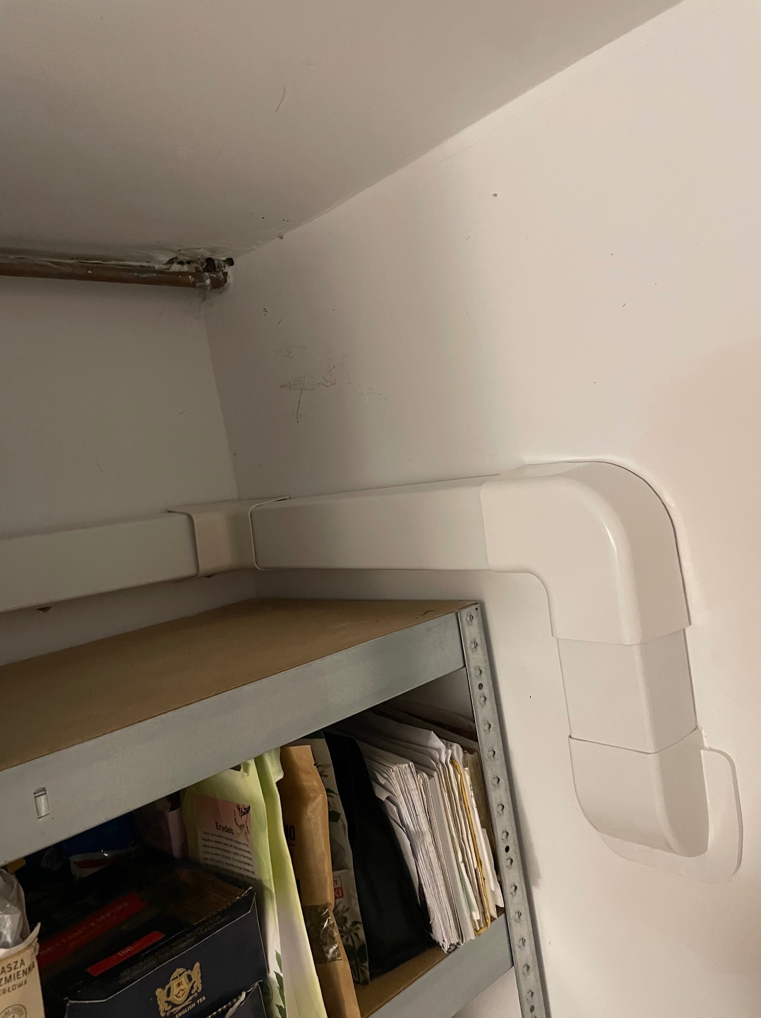 Białe rury klimatyzacji poprowadzone w rogu pomieszczenia, z metalowym regałem i przechowywanymi przedmiotami w tle.