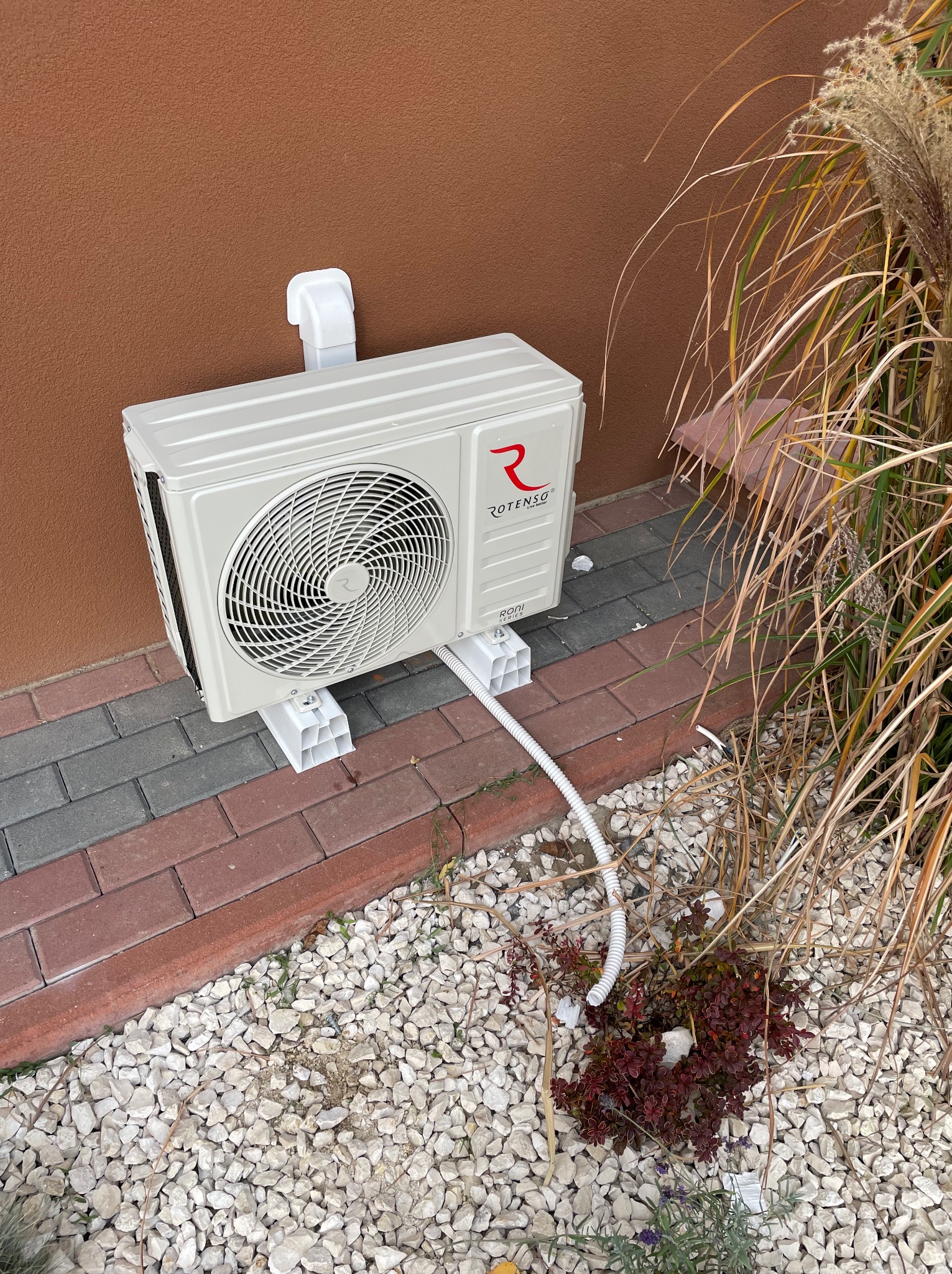 Zewnętrzna jednostka klimatyzacji Rotenso zamontowana na brązowej ścianie domu, widoczna z boku, z przewodami i elementami montażowymi na tle kamieni ozdobnych i roślinności.