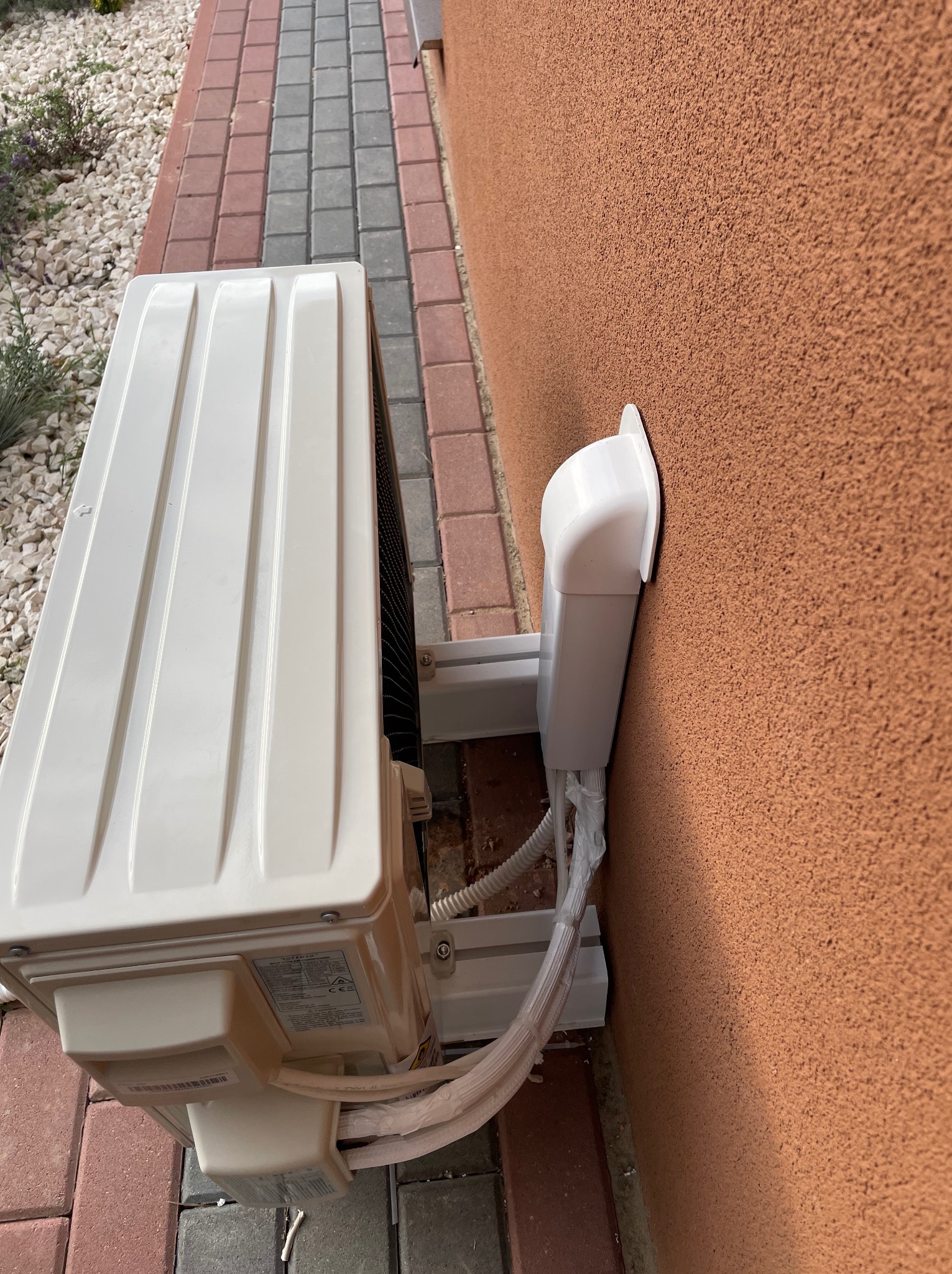 Zewnętrzna jednostka klimatyzatora zamontowana na ścianie budynku z pomarańczową elewacją, widoczne kable w plastikowej osłonie, obok kostka brukowa i kamyki.