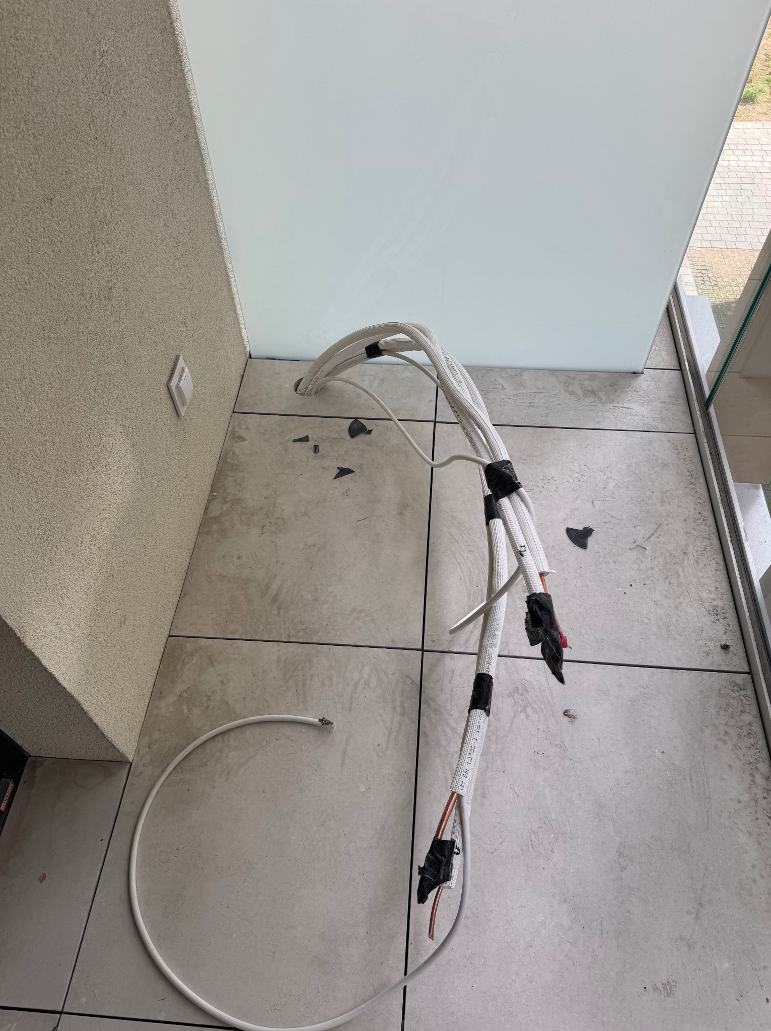 Białe przewody klimatyzacji w trakcie montażu, wychodzące z otworu w ścianie na tle płytek podłogowych i fragmentu okna. Widoczne elementy instalacji.