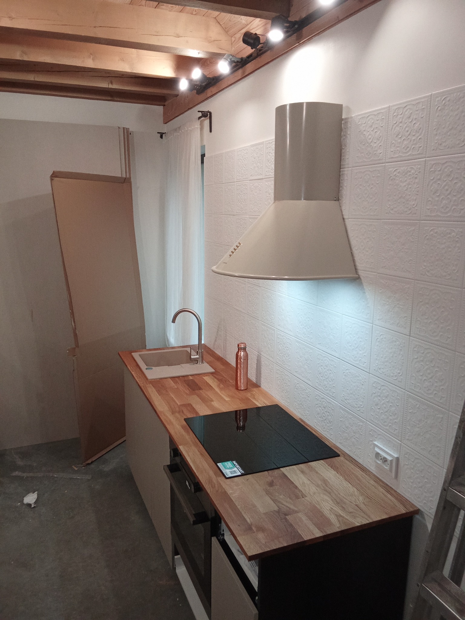 Kuchnia w trakcie montażu: drewniany blat, płyta indukcyjna, beżowy okap i ozdobne białe płytki ścienne, widoczne elementy drewnianej konstrukcji sufitu i kartonowe pudła.