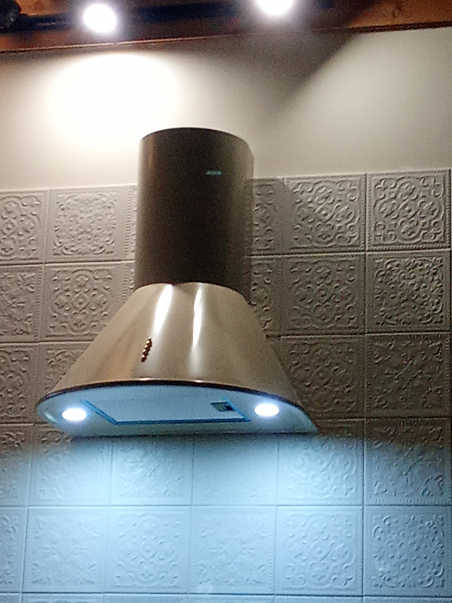 Zamontowany okap kuchenny z oświetleniem LED na tle ozdobnych, kwadratowych płytek ściennych. Widoczne elementy instalacji nad okapem.