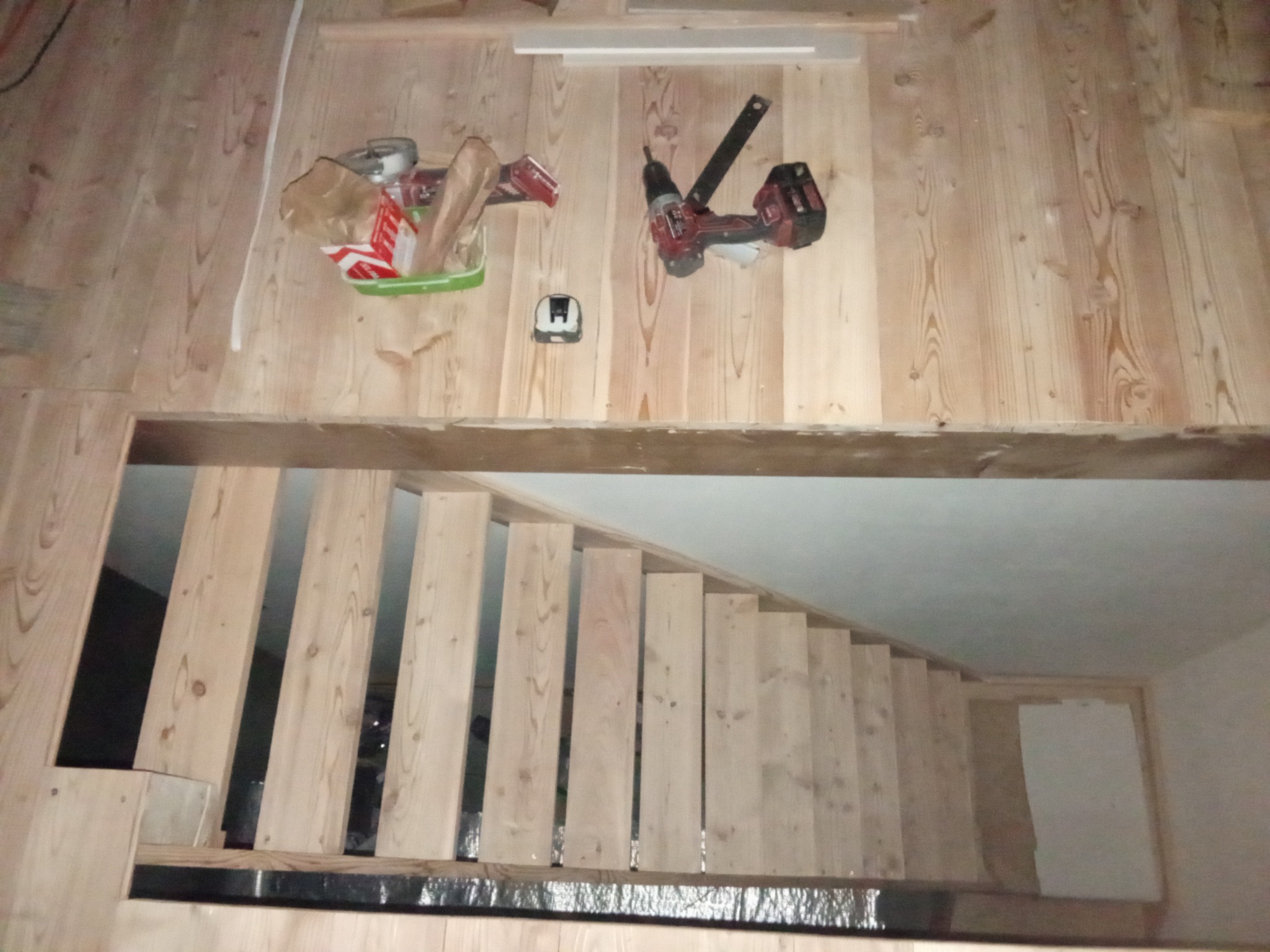 Drewniane schody w trakcie montażu, widok z góry, narzędzia i materiały budowlane na drewnianej podłodze, surowy stan wykończenia.