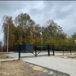 Czarna brama wjazdowa dwuskrzydłowa z ogrodzeniem panelowym na betonowej podbudowie, widok z zewnątrz na tle jesiennego krajobrazu i szarego nieba.