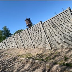 M&G Fences Konrad Smetaniuk - Długi, szary mur z prefabrykowanych elementów betonowych imitujących cegłę, ciągnący się wzdłuż ziemistej skarpy, z wieżą ciśnień w tle pod błękitnym niebem.