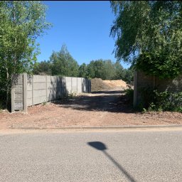 M&G Fences Konrad Smetaniuk - Fragment betonowego ogrodzenia panelowego z widocznymi słupkami imitującymi cegłę, ziemia i hałda piasku w tle, błękitne niebo