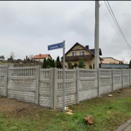 Długi, szary, betonowy płot z dekoracyjnymi elementami, widoczny z ulicy Ostrog&oacute;rskiej, z domami w tle i mężczyzną po lewej stronie.