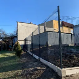 Nowoczesne ogrodzenie panelowe z podmurówką betonową na tle zabudowań, z młodym drzewkiem iglastym wzdłuż linii ogrodzenia.