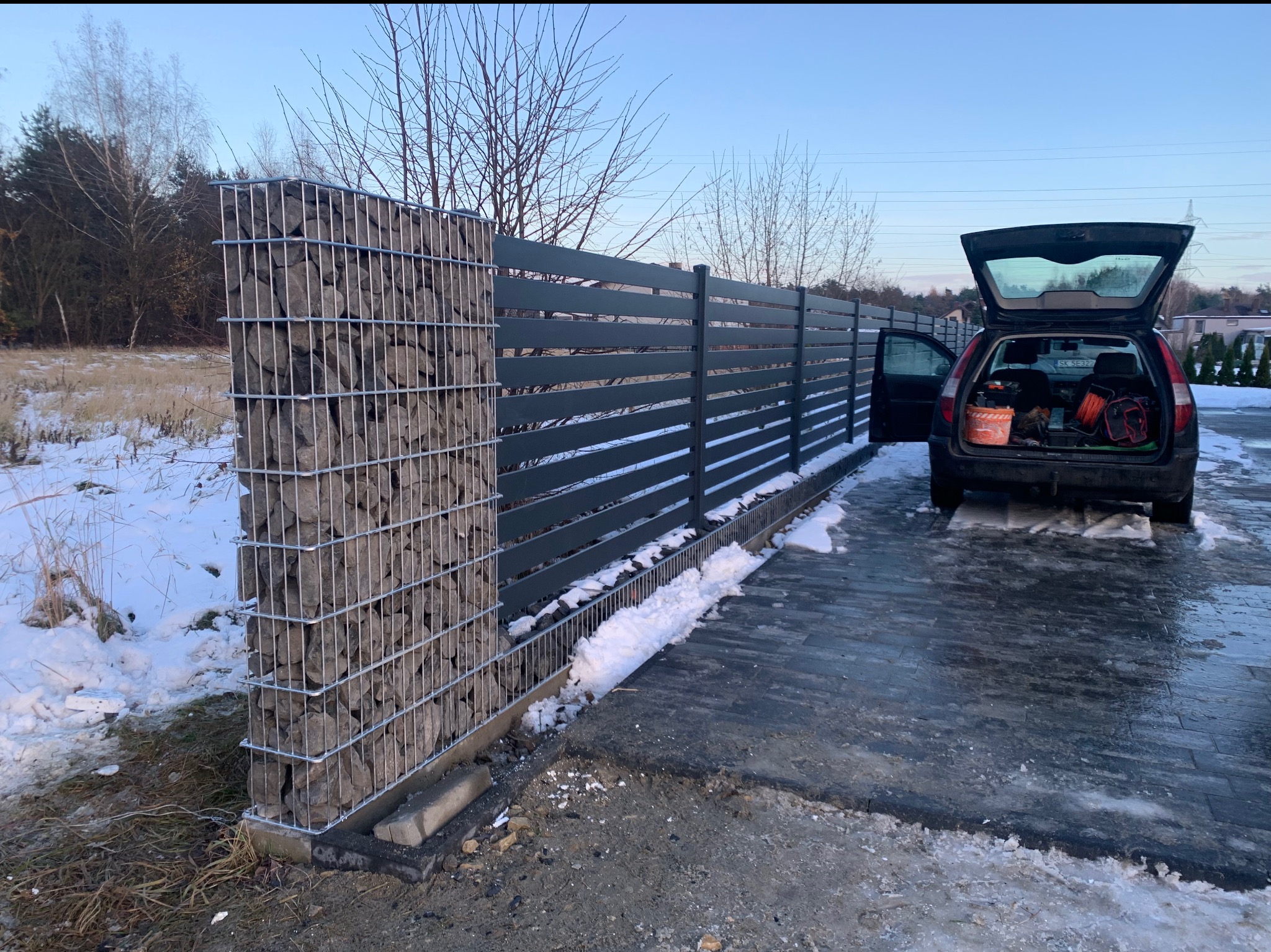 Nowoczesne ogrodzenie z gabionów i poziomych paneli w kolorze antracytowym, widok z boku z otwartym bagażnikiem samochodu z narzędziami, zimowy krajobraz z resztkami śniegu.