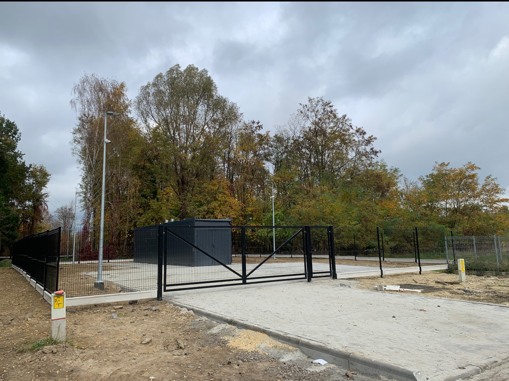 Czarna brama wjazdowa dwuskrzydłowa z ogrodzeniem panelowym na betonowej podbudowie, widok z zewnątrz na tle jesiennego krajobrazu i szarego nieba.