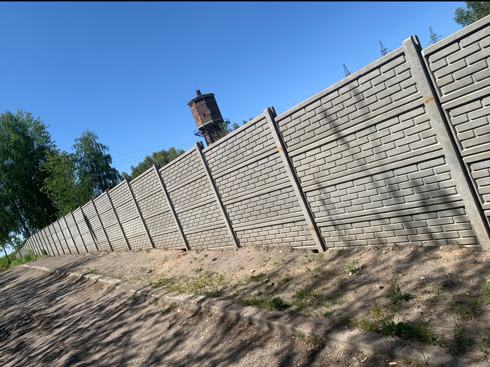 Długi, szary mur z prefabrykowanych elementów betonowych imitujących cegłę, ciągnący się wzdłuż ziemistej skarpy, z wieżą ciśnień w tle pod błękitnym niebem.