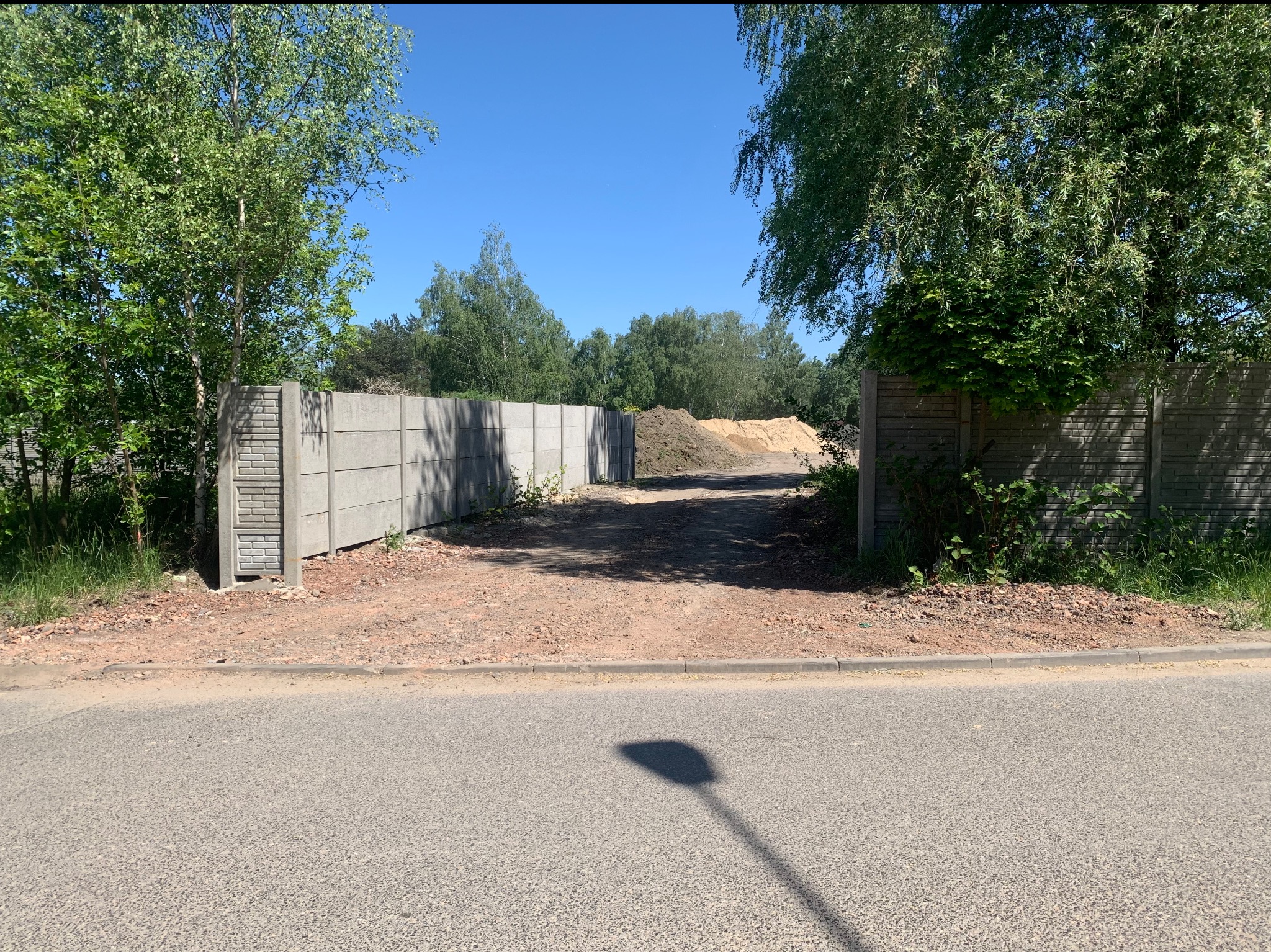 Fragment betonowego ogrodzenia panelowego z widocznymi słupkami imitującymi cegłę, ziemia i hałda piasku w tle, błękitne niebo