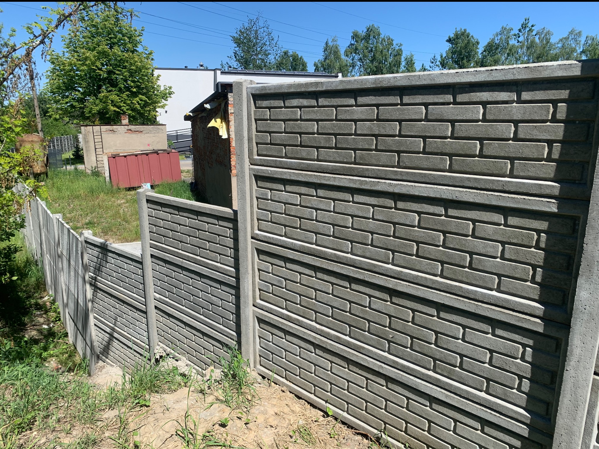 Szary betonowy płot imitujący mur z cegieł, widok z boku, z elementami trawy i zieleni w tle oraz fragmentami zabudowań.