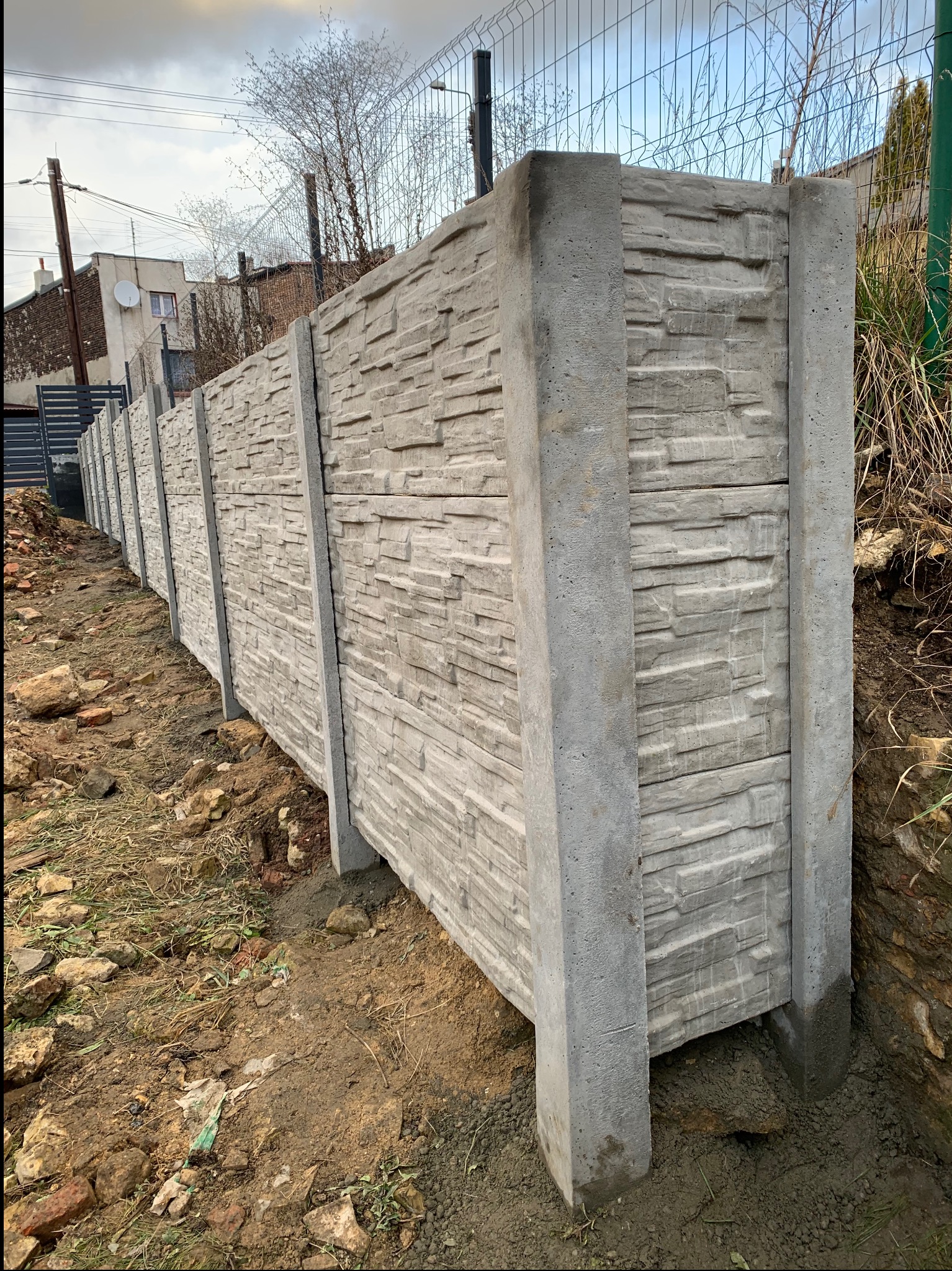 Długi, szary, betonowy mur z panelami o fakturze kamienia, widoczny z perspektywy gruntu, z ziemią i gruzem w tle.