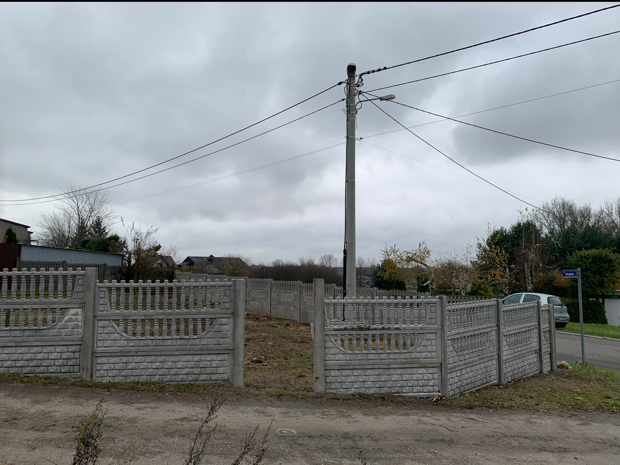 Fragment ogrodzenia betonowego w trakcie montażu, widoczne słupki i przęsła bez wypełnienia, słup energetyczny w tle, pochmurne niebo.