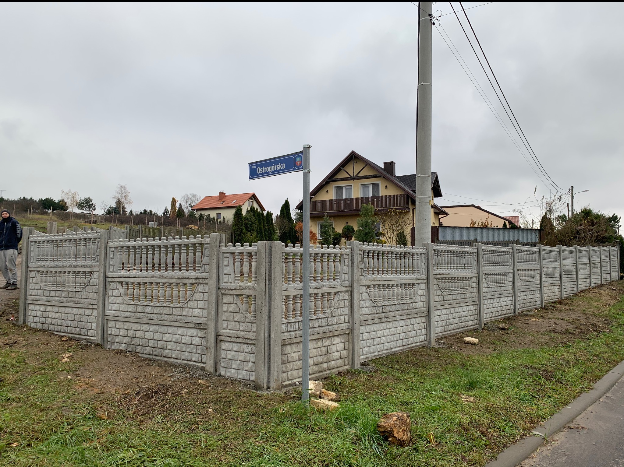 Długi, szary, betonowy płot z dekoracyjnymi elementami, widoczny z ulicy Ostrogórskiej, z domami w tle i mężczyzną po lewej stronie.