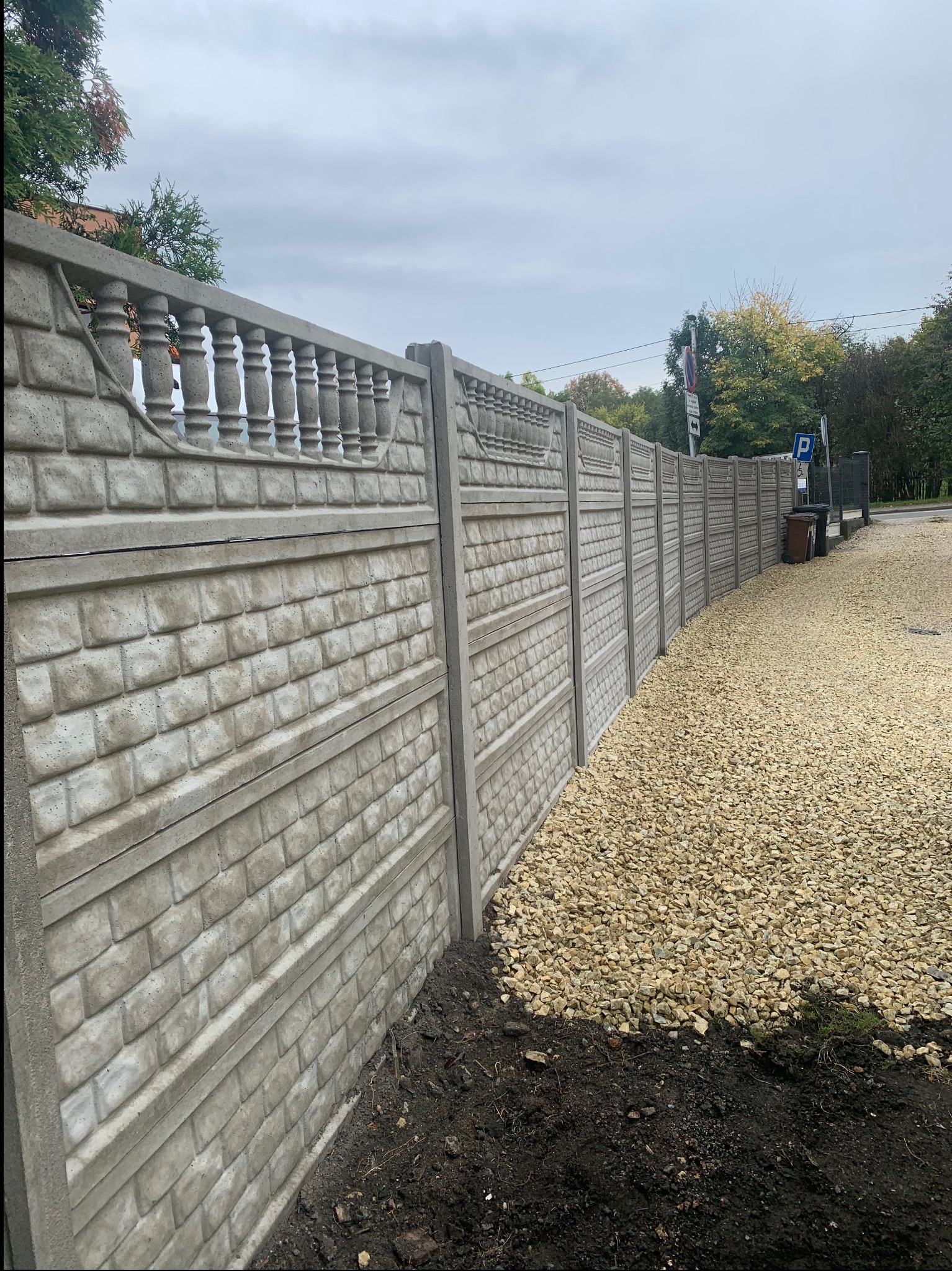Długi, szary mur z prefabrykatów betonowych imitujących cegłę, zakończony dekoracyjnym elementem z balustradą, oddzielający posesję od żwirowego parkingu z koszami na śmieci i znakiem drogowym w tle.