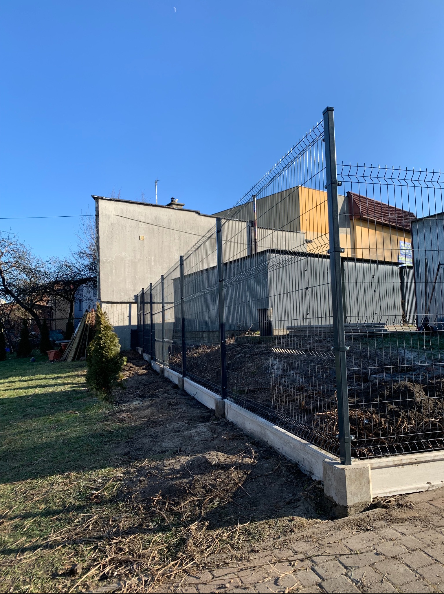 Nowoczesne ogrodzenie panelowe z podmurówką betonową na tle zabudowań, z młodym drzewkiem iglastym wzdłuż linii ogrodzenia.