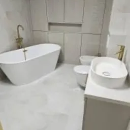 Nowoczesna łazienka z wolnostojącą białą wanną, złotą armaturą, podwieszaną toaletą i umywalką nablatową, wykończona jasnymi płytkami imitującymi kamień, z zabudową meblową pod sufit.