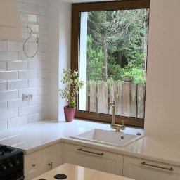 FORMAT MEBEL Henryk Rakowski - Biała kuchnia z oknem na las: Zlewozmywak z baterią w stylu retro, doniczka z rośliną. Fronty szafek frezowane z delikatnymi, złotymi uchwytami.