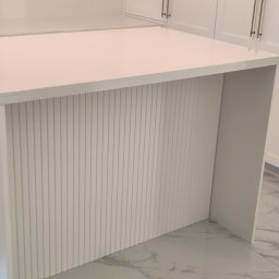 FORMAT MEBEL Henryk Rakowski - Biała wyspa kuchenna z ryflowanym frontem i blatem z konglomeratu, widoczne srebrne uchwyty szafek w tle, marmurowe płytki podłogowe.