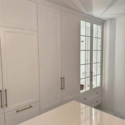 FORMAT MEBEL Henryk Rakowski - Biała zabudowa kuchenna z przeszkloną witryną i złotymi uchwytami, uzupełniona o blat. Minimalistyczny design, subtelne oświetlenie, marmurowa podłoga.