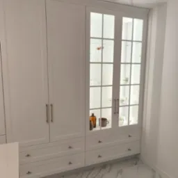 Biała zabudowa meblowa z przeszkloną witryną, podświetloną od wewnątrz. Widoczne naczynia i dekoracje. Marmurowa podłoga. Ujęcie z dołu.