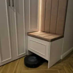 Hall z siedziskiem i szafą w zabudowie, oświetlony listwą LED. Robot sprzątający pod siedziskiem. Klasyczne frezy na frontach szafy i siedziska. Parkiet ułożony w jodełkę.