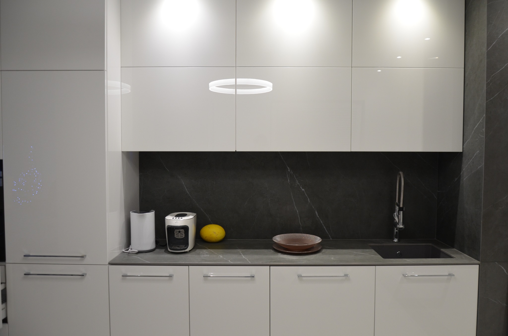 Nowoczesna kuchnia z białymi, lakierowanymi szafkami, blatem w odcieniach szarości i czarnym panelem ściennym imitującym marmur; okrągła lampa sufitowa, ekspres do kawy, owoc i naczynia na blacie.