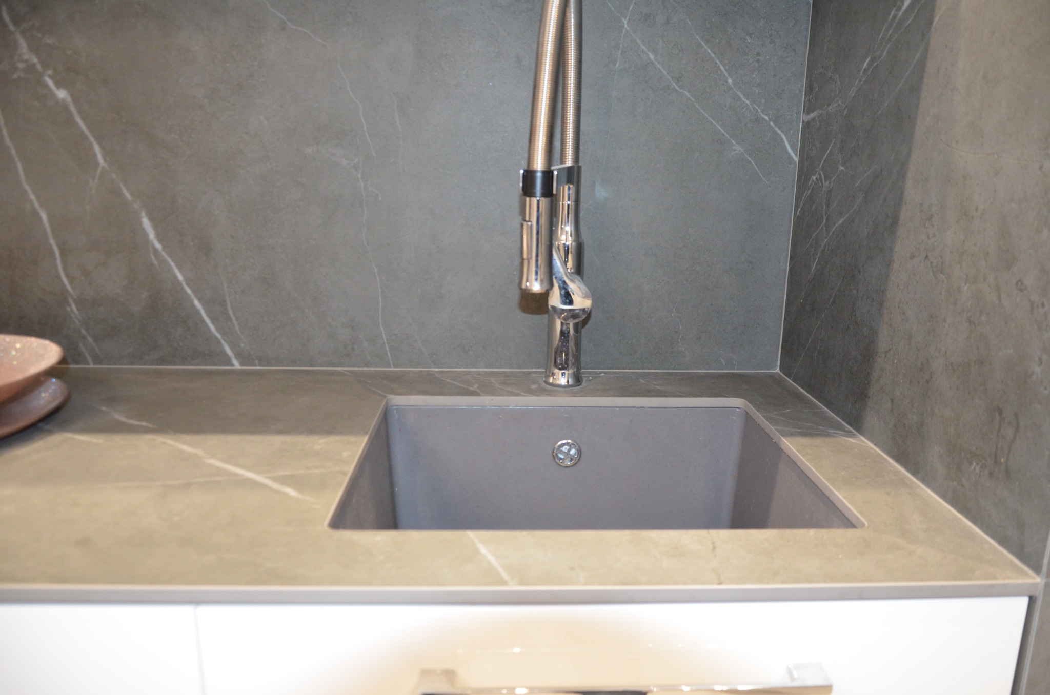 Szary zlew z baterią z wyciąganą wylewką, wkomponowany w blat z delikatnym marmurkowym wzorem, na tle ściany wyłożonej płytkami imitującymi marmur.