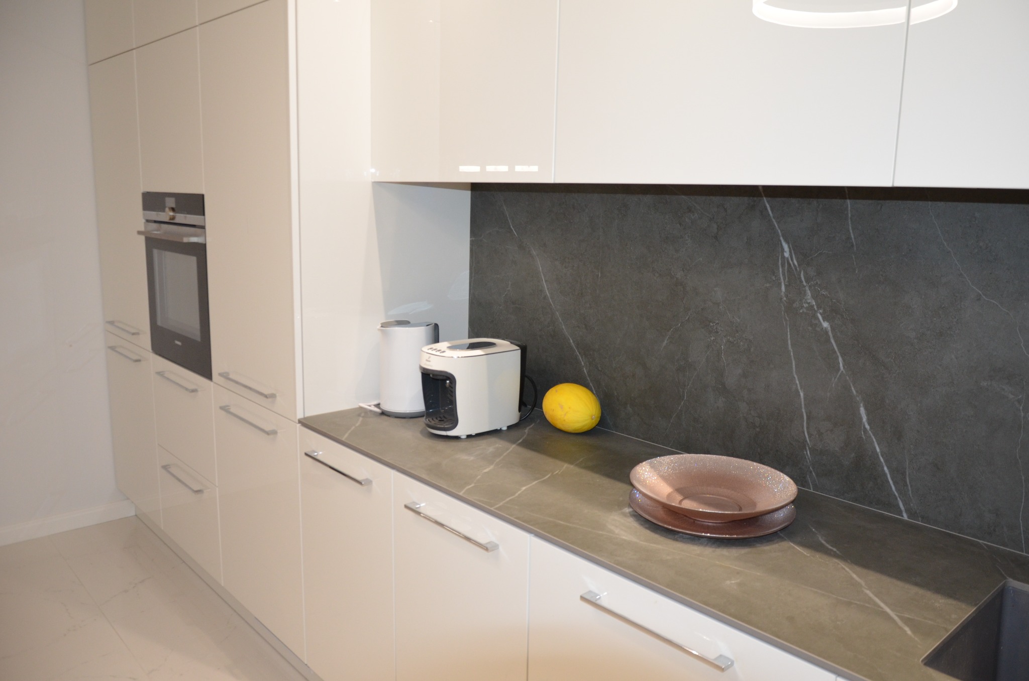 Biała, nowoczesna kuchnia z błyszczącymi frontami szafek, szarym blatem imitującym marmur i sprzętami AGD: piekarnikiem w zabudowie, czajnikiem i ekspresem do kawy. Na blacie dekoracyjna misa...