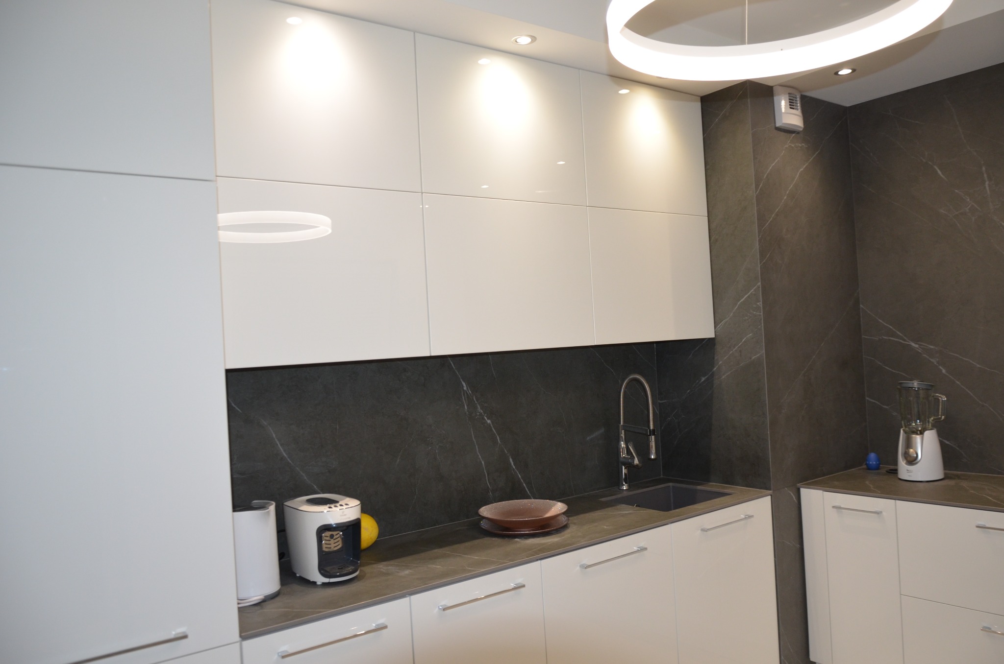 Nowoczesna kuchnia z białymi, lakierowanymi frontami, ciemnym blatem i marmurowym backsplashiem, oświetlona okrągłą lampą sufitową LED i punktowymi reflektorami.
