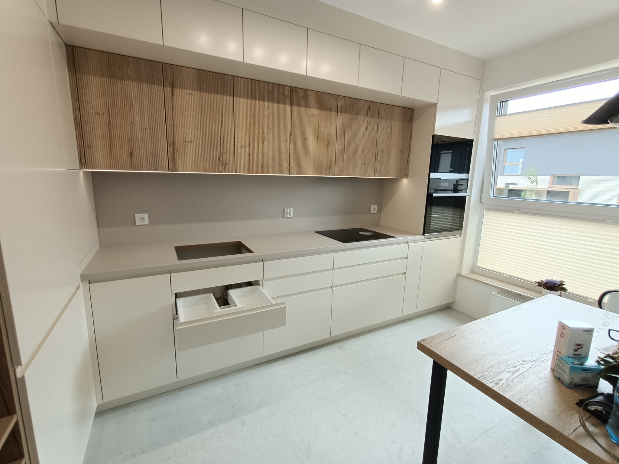 Nowoczesna kuchnia na wymiar z białymi szafkami dolnymi i górnymi w kolorze drewna, otwartą szufladą z organizerem, czarną płytą grzewczą i piekarnikiem w zabudowie.