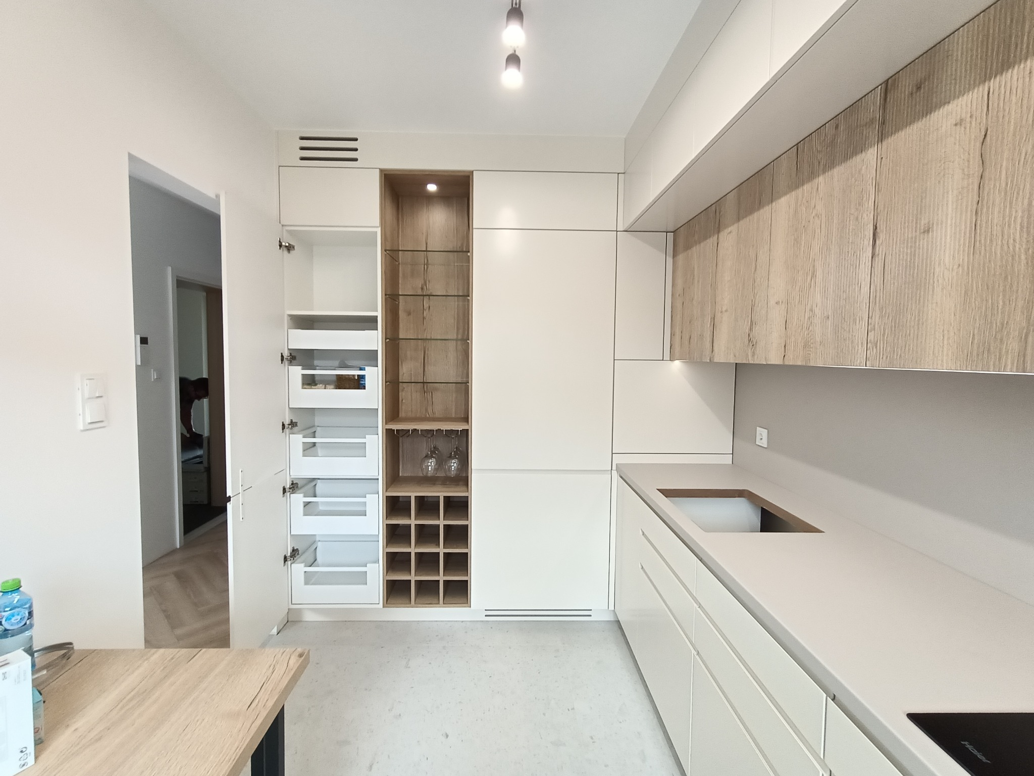 Nowoczesna kuchnia z białymi frontami i elementami drewnopodobnymi, widoczne otwarte szafki cargo, szklana witryna z miejscem na wino i kieliszki, minimalistyczny blat z wyciętym otworem na zlew.