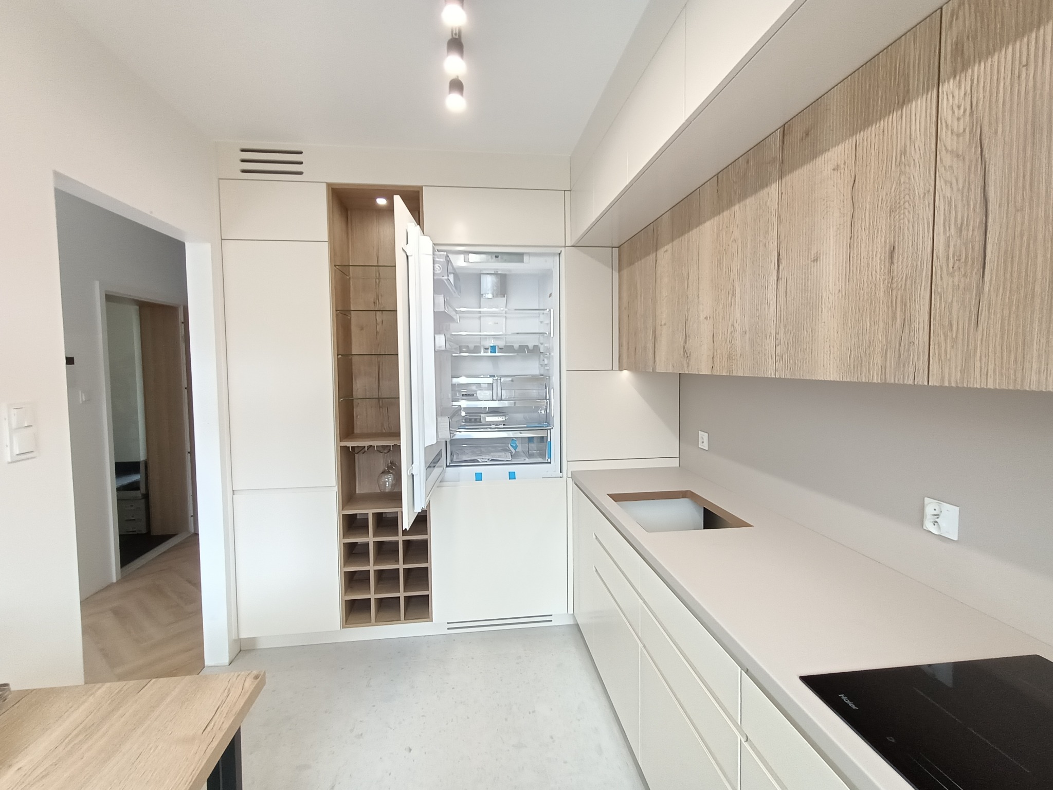 Nowoczesna kuchnia z białymi frontami i drewnopodobnymi szafkami wiszącymi, z otwartą lodówką w zabudowie i regałem na wino, widok na parkiet w jodełkę w przejściu do innego pomieszczenia.