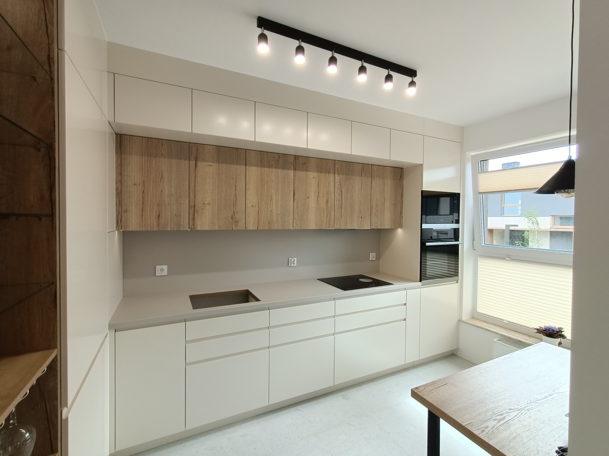 Jasna kuchnia z drewnianymi akcentami: fronty szafek górnych z wyraźnym rysunkiem drewna, płyta indukcyjna na blacie, oświetlenie punktowe na szynie sufitowej, widok na okno z żaluzjami plisowanymi.