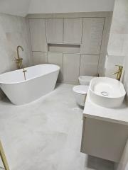 Nowoczesna łazienka z wolnostojącą białą wanną, złotą armaturą, podwieszaną toaletą i umywalką nablatową, wykończona jasnymi płytkami imitującymi kamień, z zabudową meblową pod sufit.