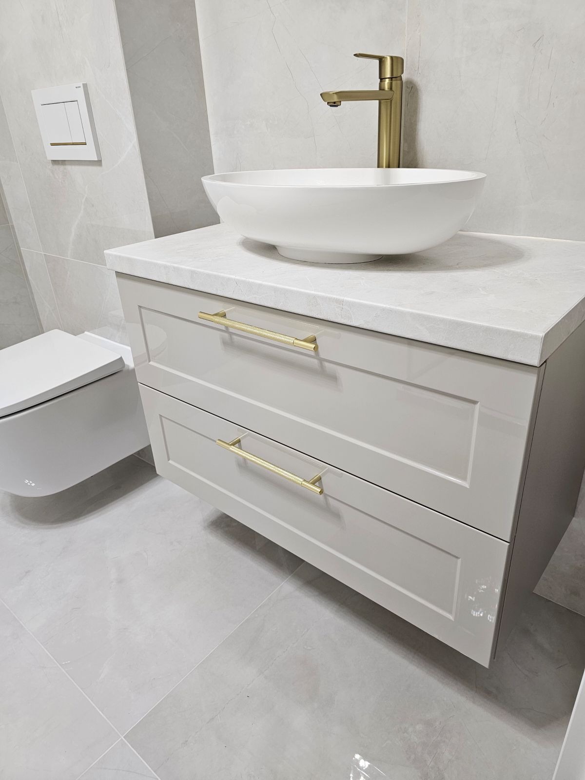Nowoczesna łazienka: biała umywalka nablatowa z złotą armaturą na szarej szafce z dwoma szufladami i złotymi uchwytami, obok podwieszana toaleta i marmurowe płytki na ścianach i podłodze.