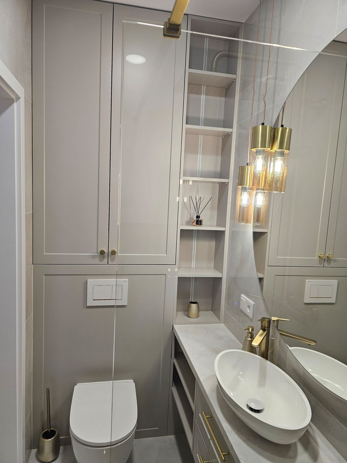 Nowoczesna łazienka w odcieniach szarości z zabudową meblową, wiszącą toaletą, umywalką nablatową i złotymi akcentami w postaci armatury i lamp wiszących.