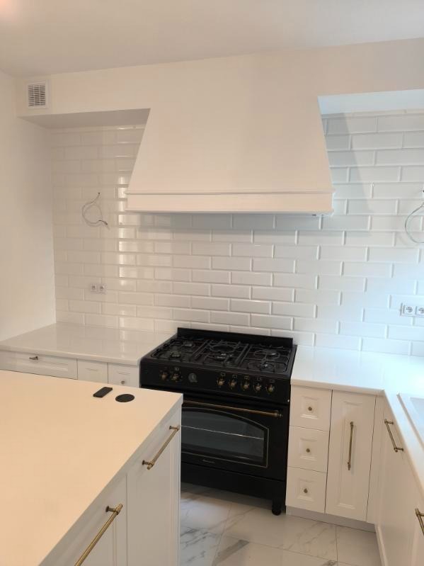 Biała kuchnia z czarną kuchenką retro i białymi, błyszczącymi płytkami na ścianie. Wyspa kuchenna z blatem w jasnym odcieniu. Złote uchwyty dodają elegancji.