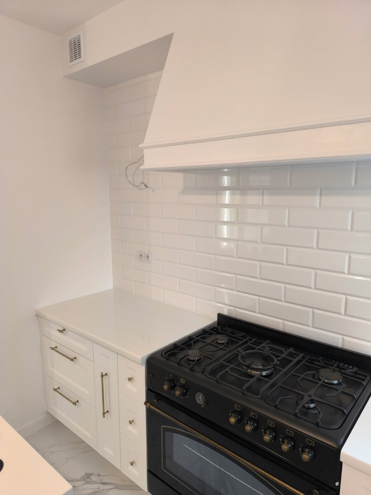 Biała kuchnia z czarną, retro kuchenką gazową i białymi płytkami typu metro. Widoczne białe szafki z złotymi uchwytami i biały blat. Minimalistyczny design z akcentem retro.