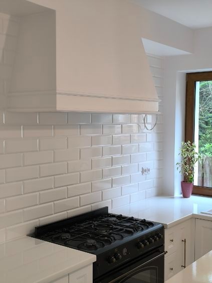 Biała kuchnia z czarną kuchenką gazową i okapem. Ściany wyłożone białymi, prostokątnymi płytkami, okno z rośliną doniczkową w tle.