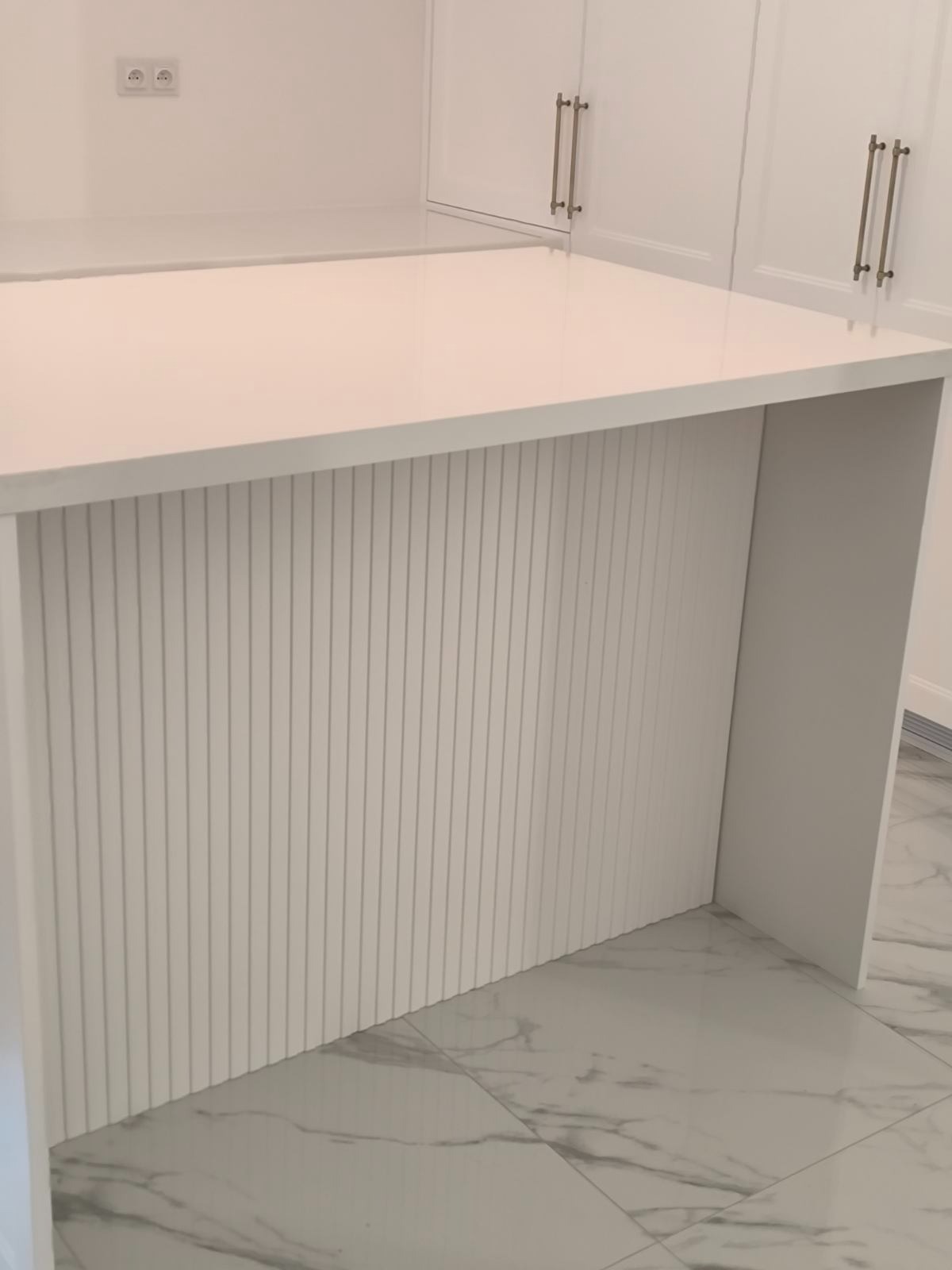 Biała wyspa kuchenna z ryflowanym frontem i blatem z konglomeratu, widoczne srebrne uchwyty szafek w tle, marmurowe płytki podłogowe.